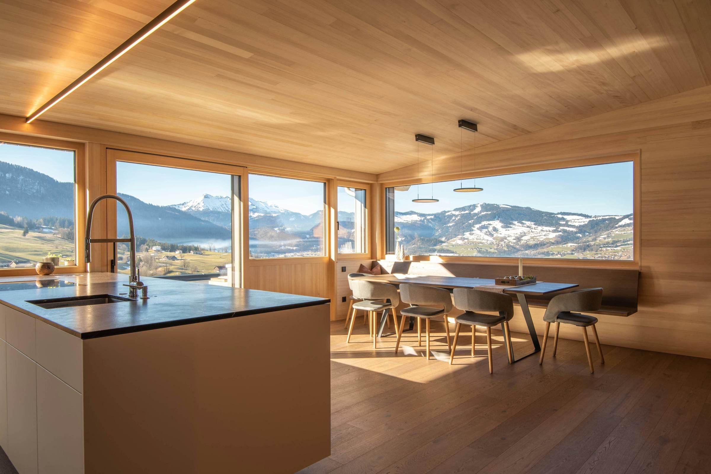 Îlot de cuisine blanc et table en bois devant une baie d’angle, soleil bas d’hiver sur le plancher en chêne