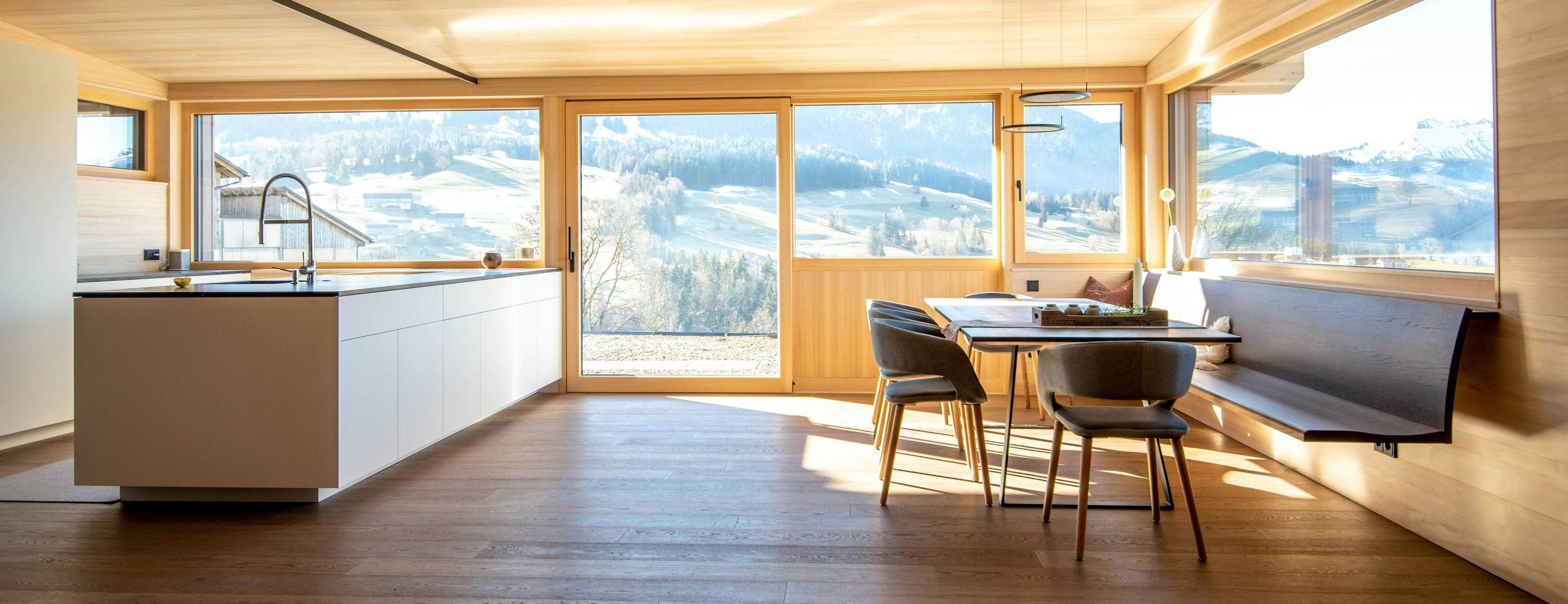 Séjour et cuisine d’un chalet rénové, plafond et murs en bois clair, grandes baies sur les pâturages enneigés et la montagne