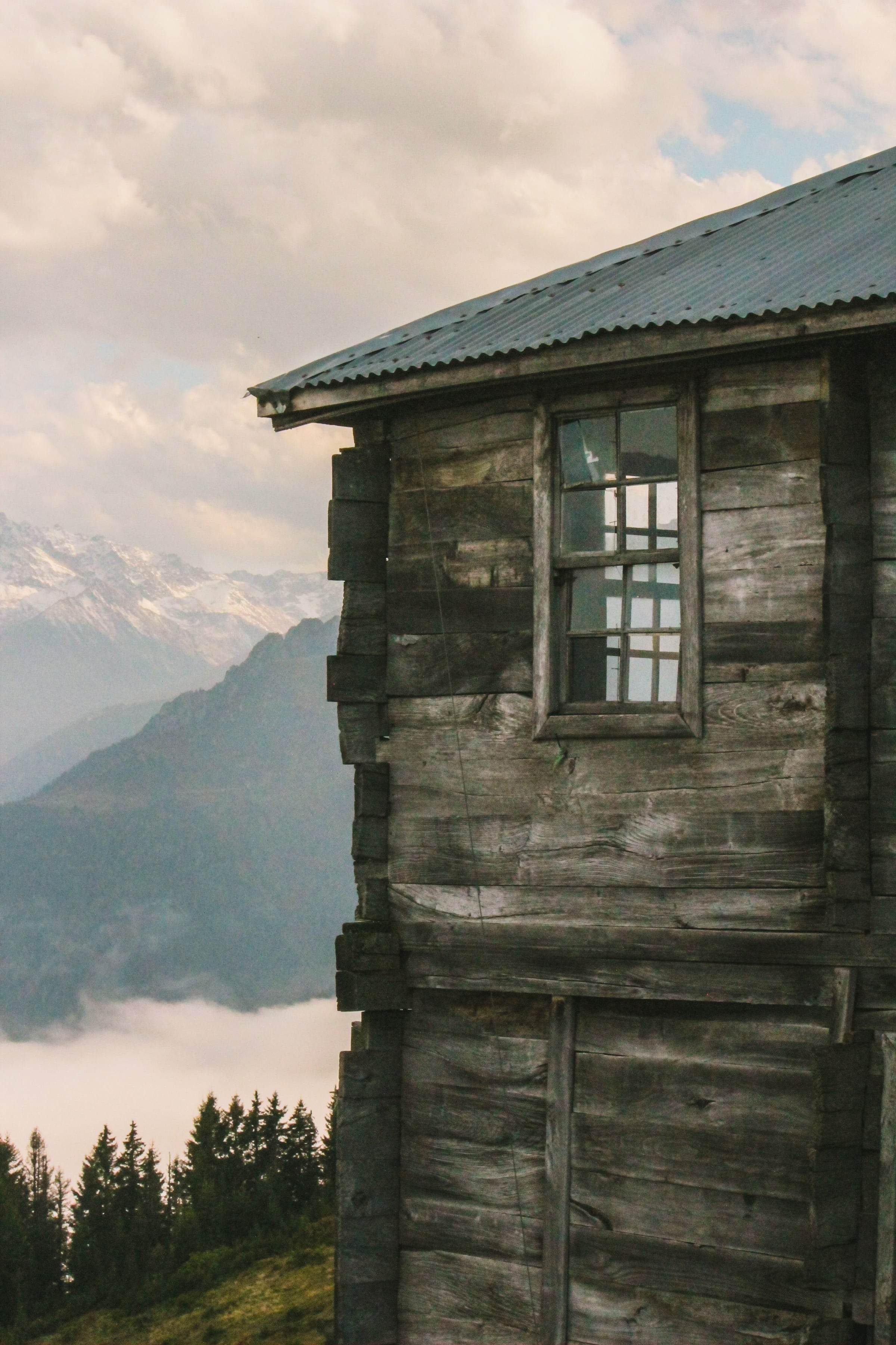 Angle d’un vieux bâtiment en madriers gris, petite fenêtre à croisillons, vallée et sommets dans la brume derrière